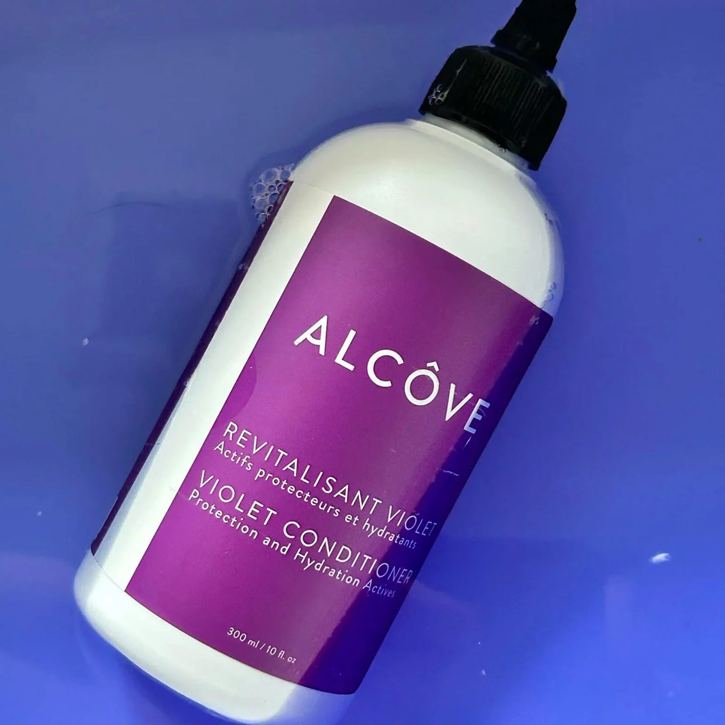 A bottle of Alcôve Revitalisant Violet / Violet Conditioner, featuring a white bottle with a prominent purple label and a black cap, set against a solid blue background.
