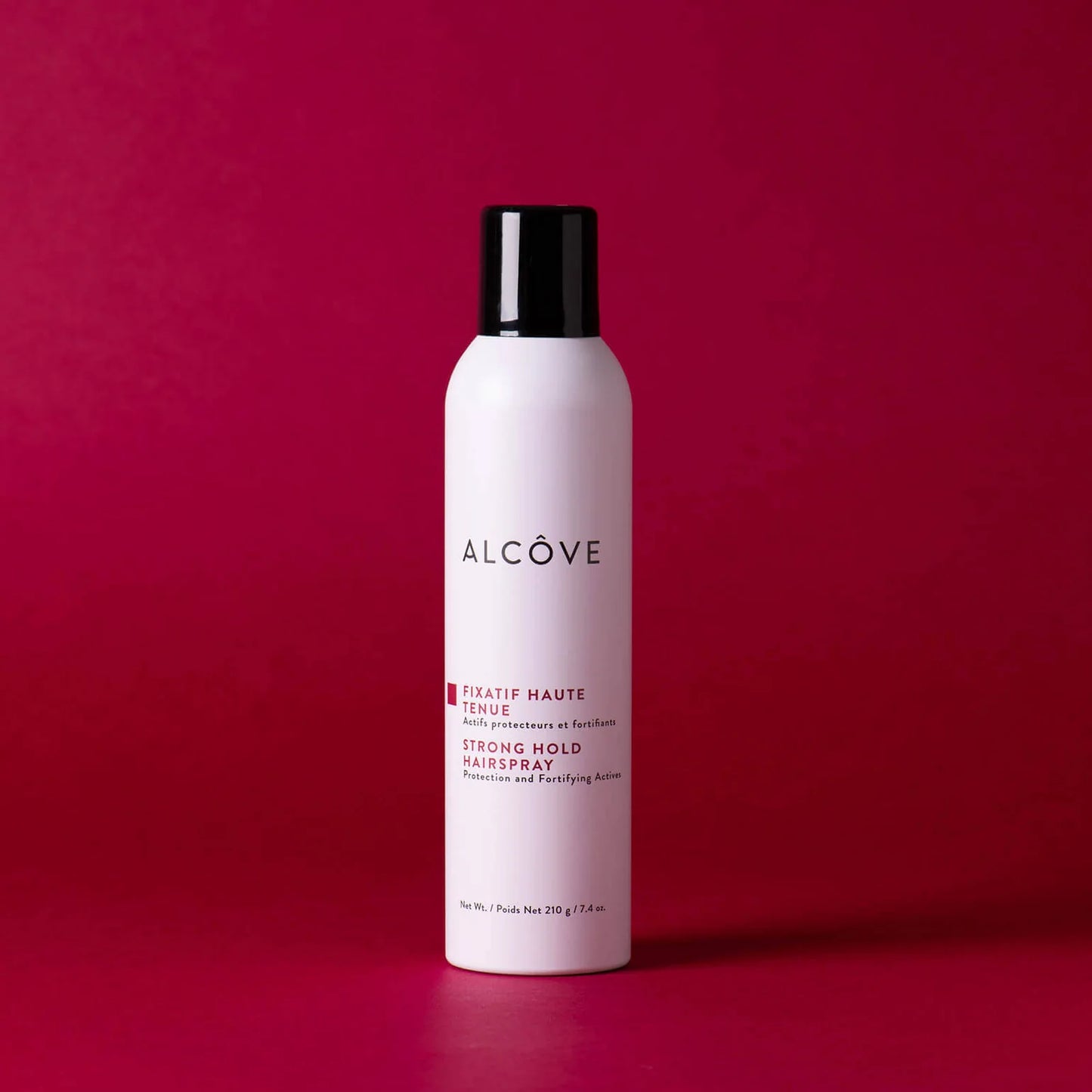 A bottle of Alcôve Strong Hold Hair Spray in a matte white bottle with a black cap, set against a solid dark magenta background.