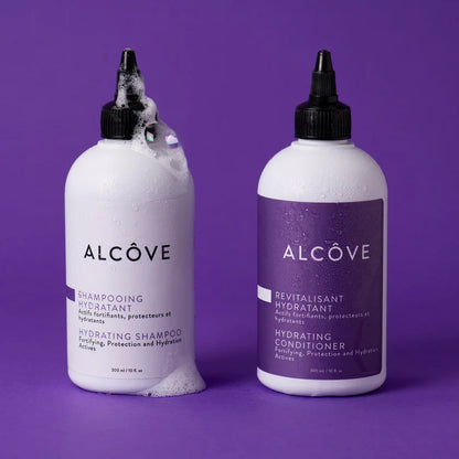 Two white plastic bottles of Alcôve haircare products, one labeled hydrating shampoo with white foam at the top and bottom, and one labeled hydrating conditioner with a purple label, set against a solid purple background.