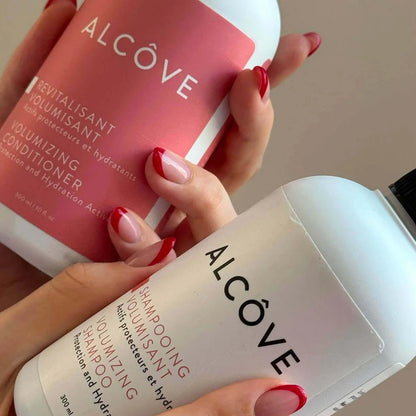 Two bottles of Alcove hair care products held by a person with red nail polish.