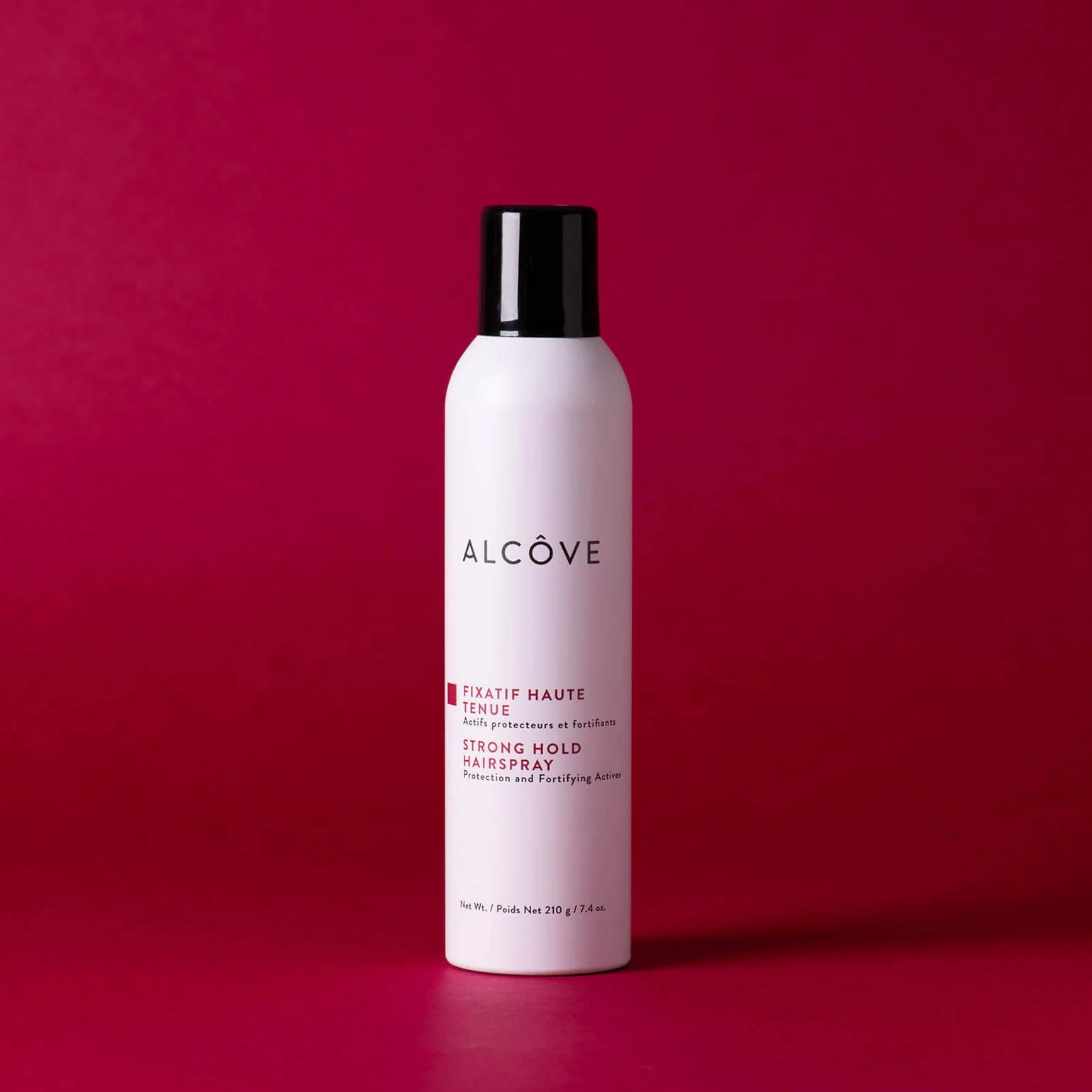 A bottle of Alcôve Strong Hold Hair Spray in a matte white bottle with a black cap, set against a solid dark magenta background.