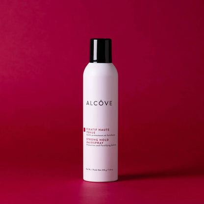 A bottle of Alcôve Strong Hold Hair Spray in a matte white bottle with a black cap, set against a solid dark magenta background.