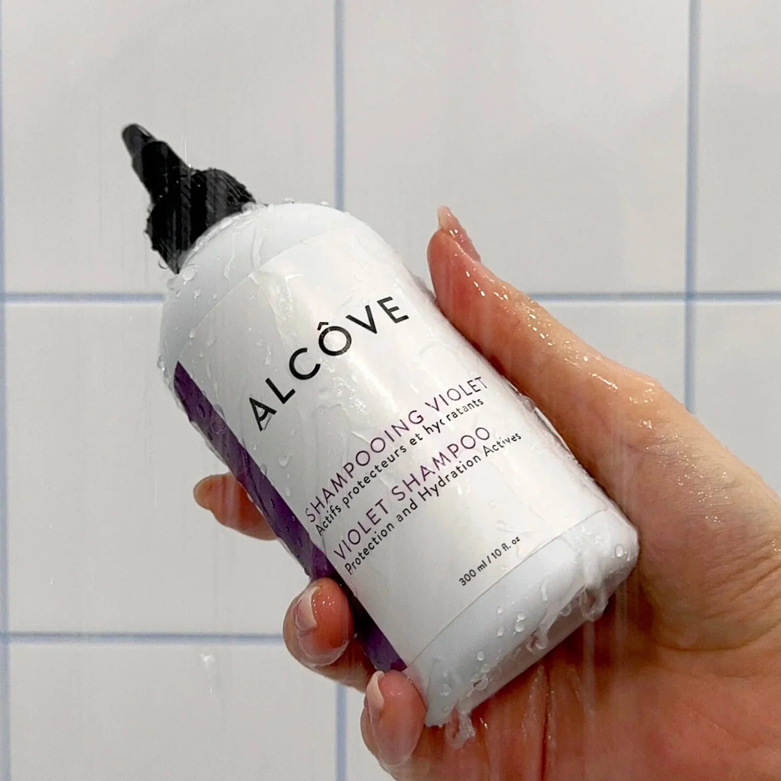 Hand holding a bottle of Alcove Violet Shampoo against a tiled wall background